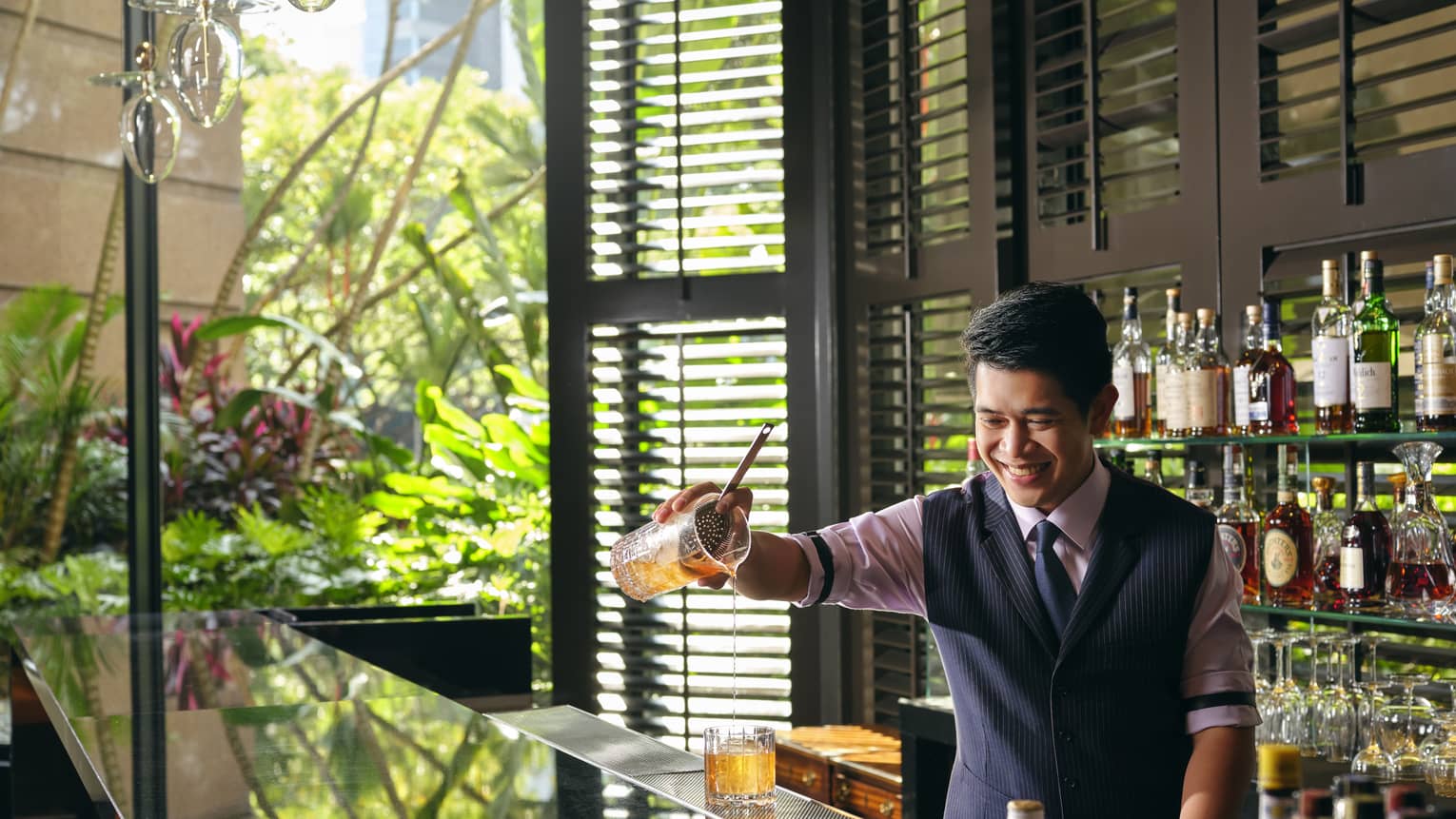 A smiling bartender pours a Sidecar cocktail, a display of liquor bottles behind him, light streaming in from the windows to his right 