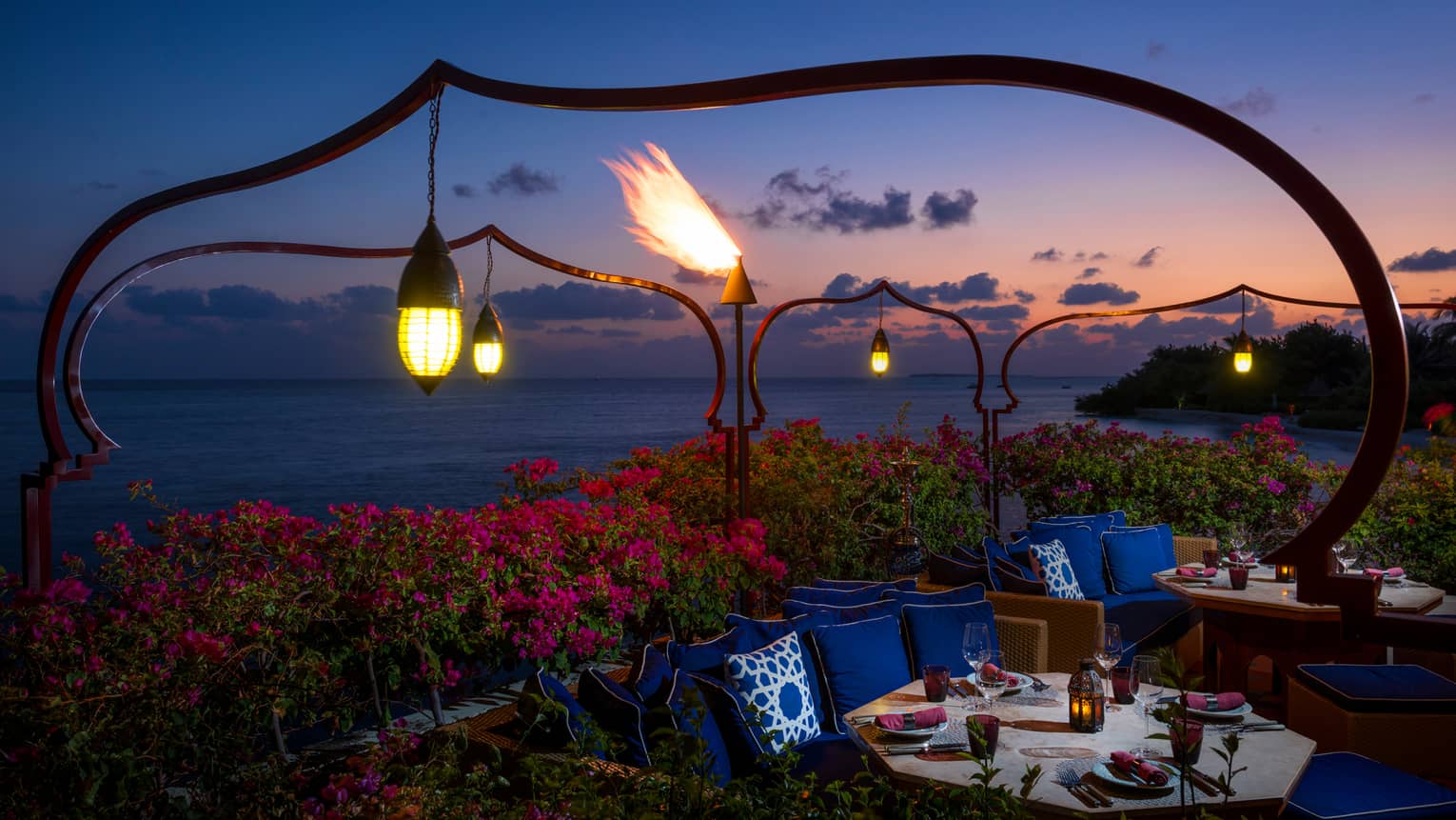 Lanterns hang over pink tropical gardens, booths with dining tables, plush blue pillows at night
