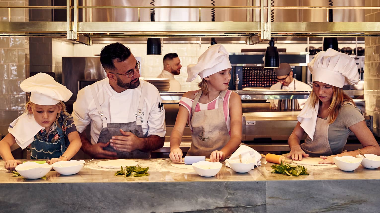 A chef standing at a counter teaching children wearing aprons and chef hats how to roll out pizza dough using rolling pins.