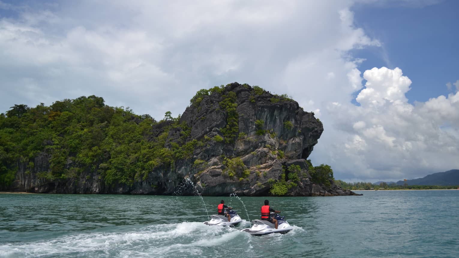 Two people wearing red life vests on jet skis ride on ocean under mountain