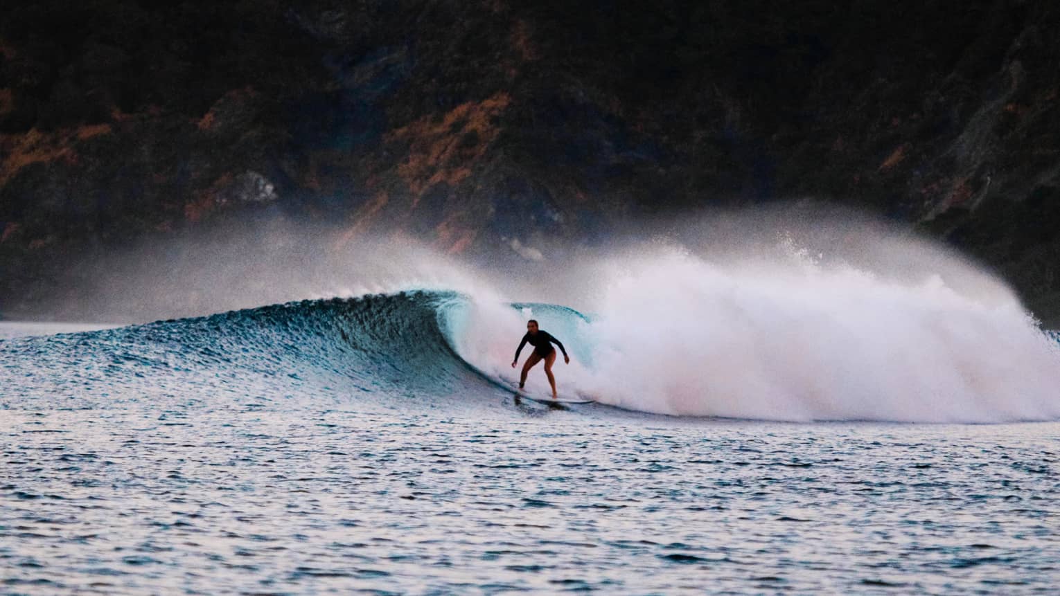 A surfer rides a wave amid a great cloud of frothy sea foam; a jagged cliff in the background, pink and yellow skies above.  