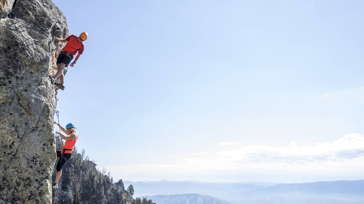 Man and woman climb steep cliff face with view of mountains in far distance 