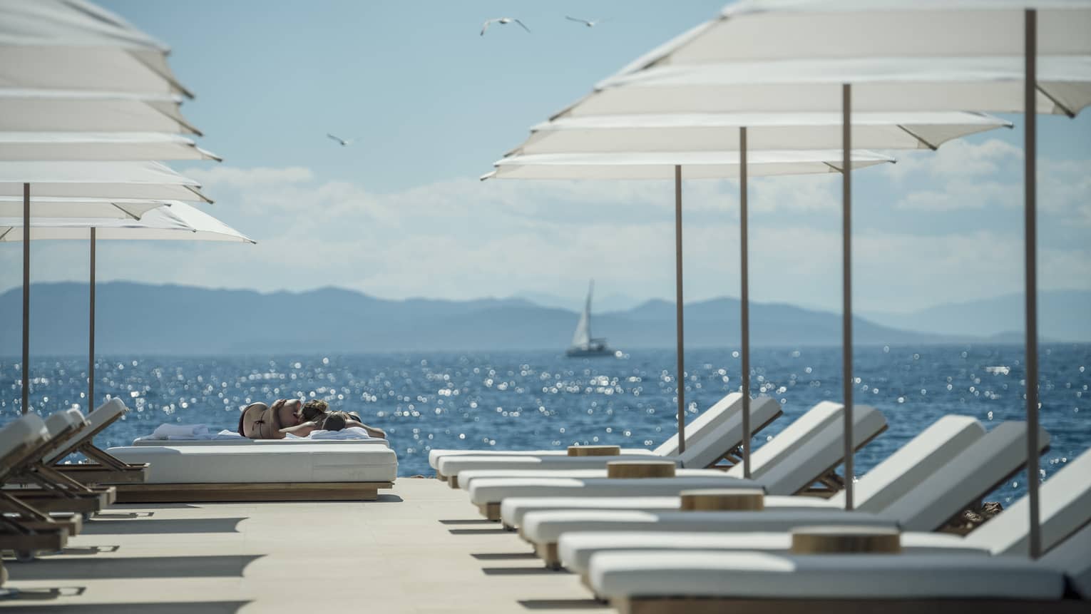 A sail boat sails in the distance while white lounge chairs are ready for guests and lined up next to the oceanside Nafsika Pool in Athens. 