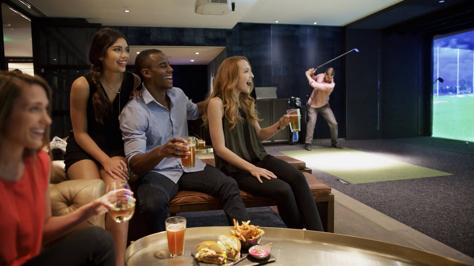 A man and three woman sip beer and cheer on a sports team in the foreground, while another man golfs in the background of a function room at four seasons hotel houston 