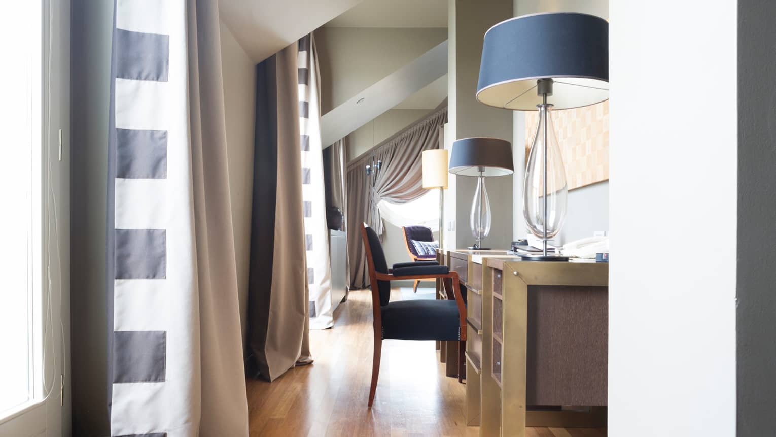 Hotel suite neutral desk topped with two glass lamps, windows behind flanked by striped curtains