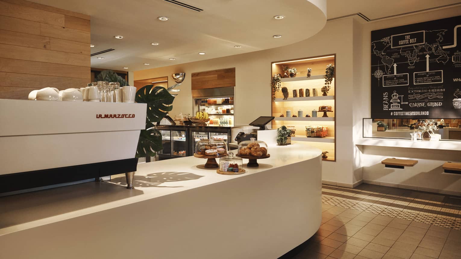 Curved white counter with a white espresso machine on top and a small display of pastries, with another display and shelving on the other side of the room
