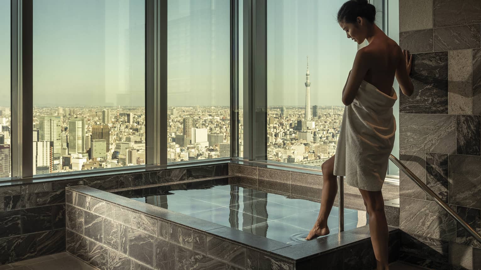 Woman wrapped in white towel dips her toe into the ofuro bath