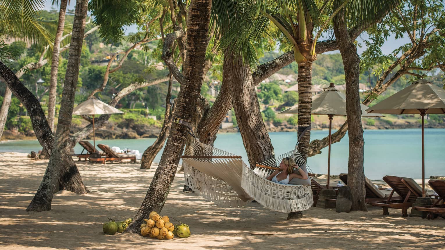 Woman in white bikini lies on white hammock in shade of large trees on Coconut Grove Beach