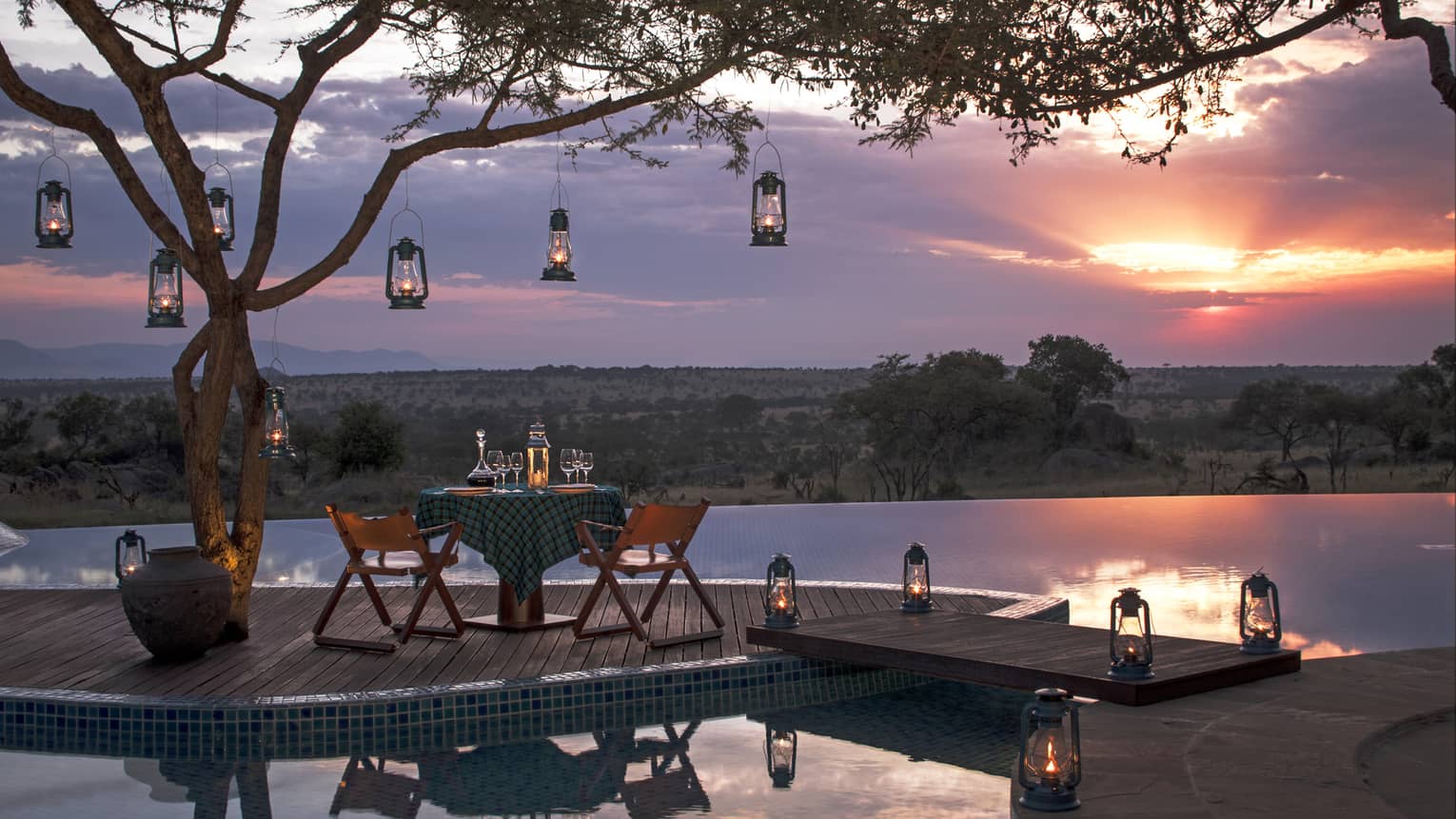 Private dining table on wood dock lined with lanterns, hanging from tree, sunset
