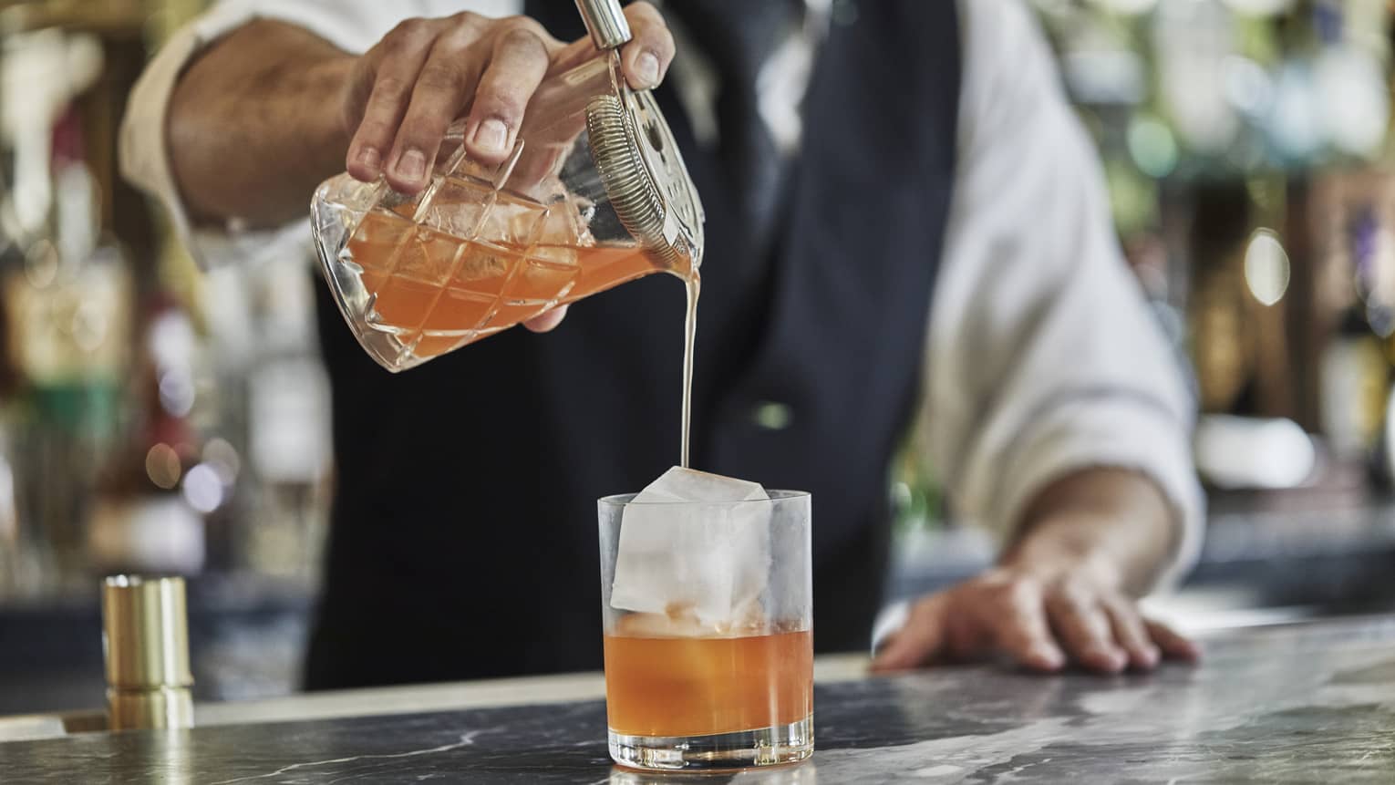 Close-up of bartender pouring orange cocktail from shaker into crystal glass with large ice cube
