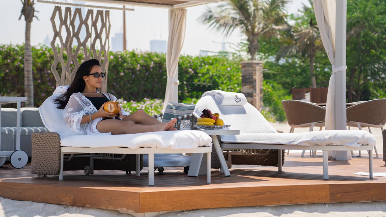 Woman in sunglasses and bathing suit lounges on chaise under poolside private cabana