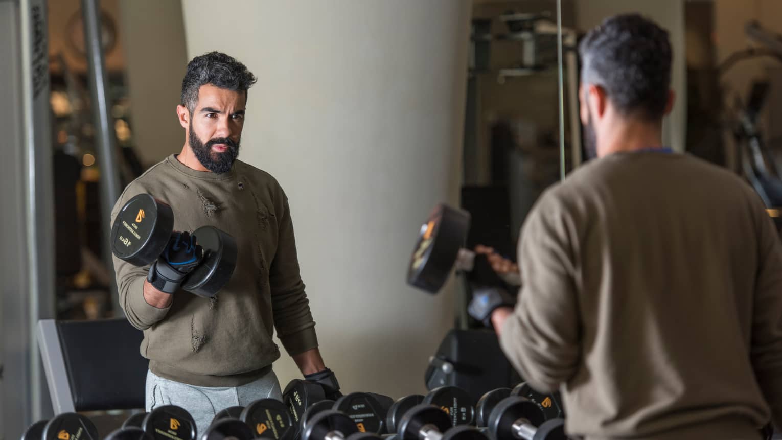 Man with beard wearing casual sweatshirt and sweatpants stands looks in gym mirror, lifts large hand weights