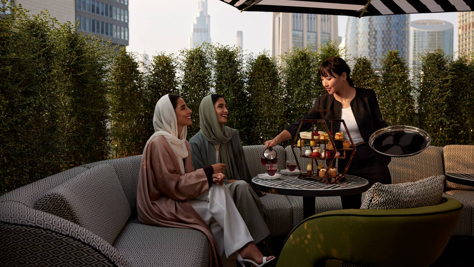 Two women sit on a beige patterned sofa beneath a black-and-white patio umbrella as a waitress serves them tea and pastries