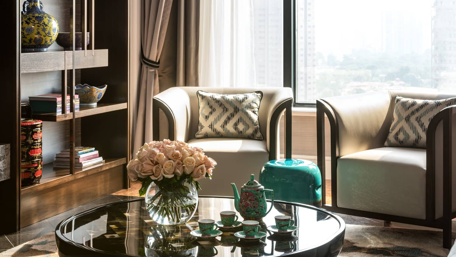 A cosy seating area with two armchairs, round coffee table holding a vase of pink roses and tea set, with a bookshelf displaying decorative vases and books nearby.