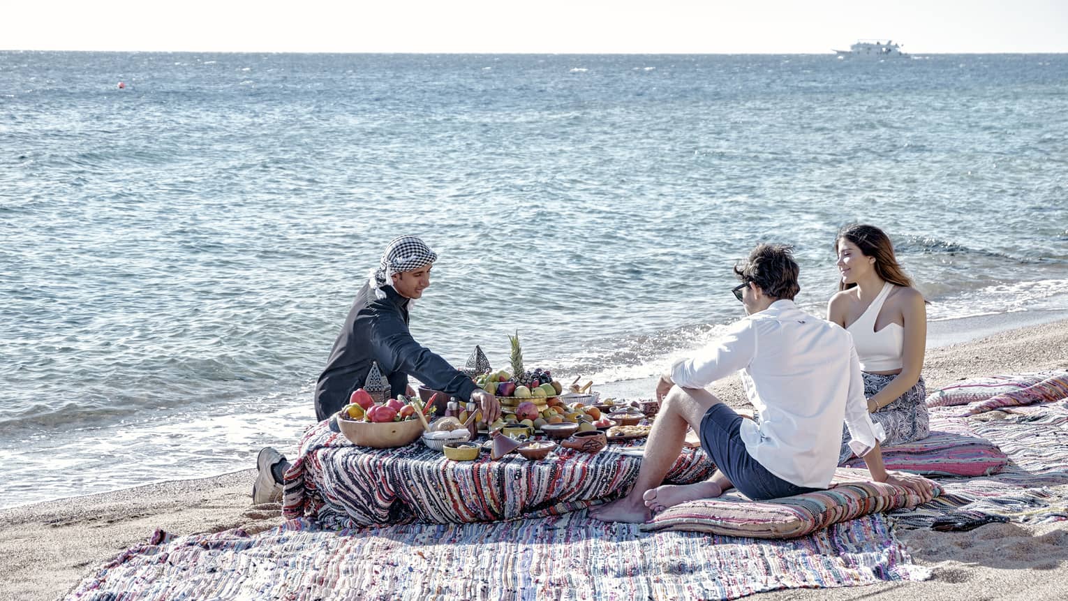 Three people sit on a beach around a low table set with a luxurious array of fruit, bread, and clay dishes full of food.