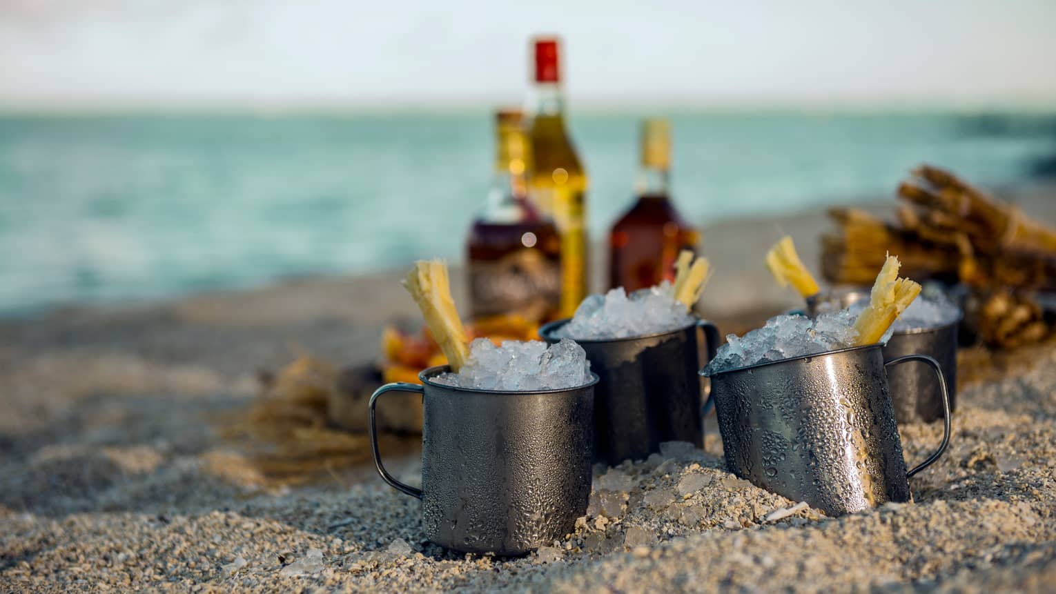 Steel cups with crushed ice in front of liquor bottles in sand on beach