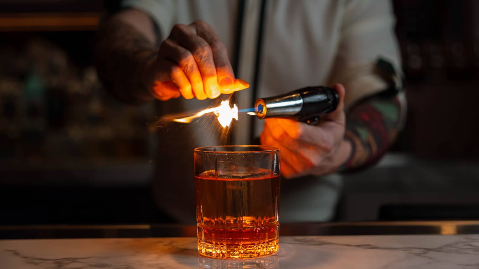Bartender using a torch to flame an orange peel over a glass of whiskey, creating a dramatic effect at the bar.