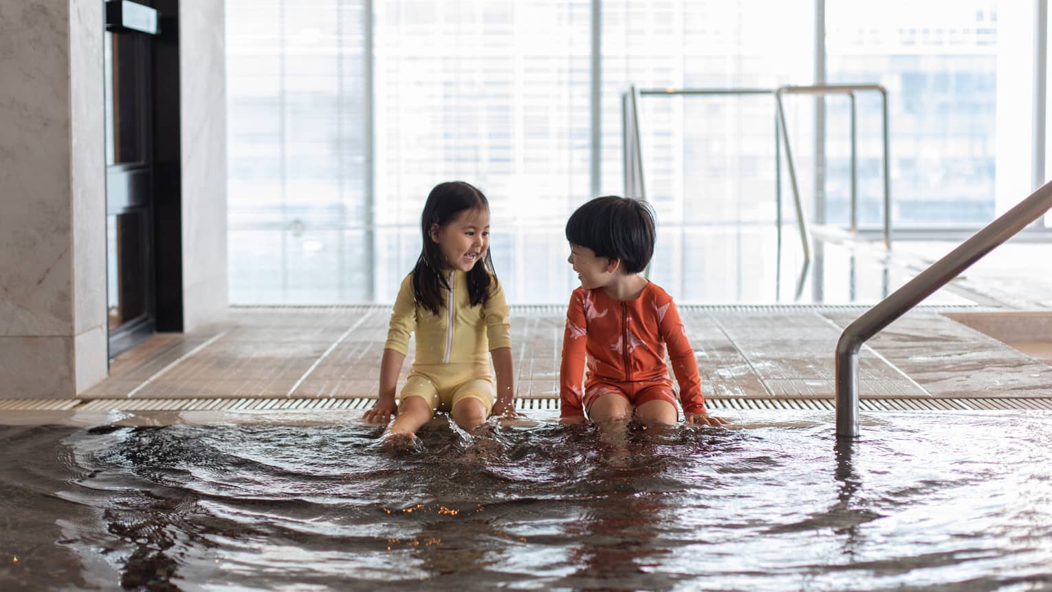 Two young laughing children sit at the edge of an indoor pool