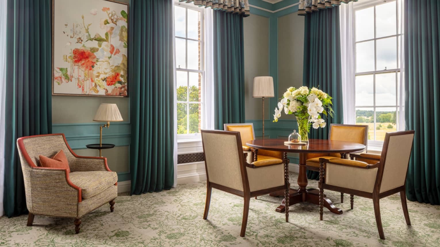 Guest room with round table and four dining chairs, tan accent chair, twin windows with blue drapes