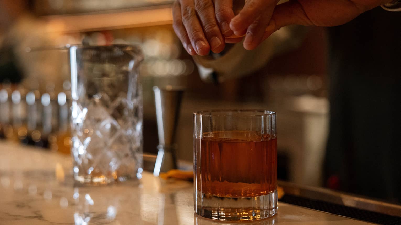 Two hands twist an orange peel over a lowball glass tumbler filled with clear brown liqueur on a smooth, marble bar.
