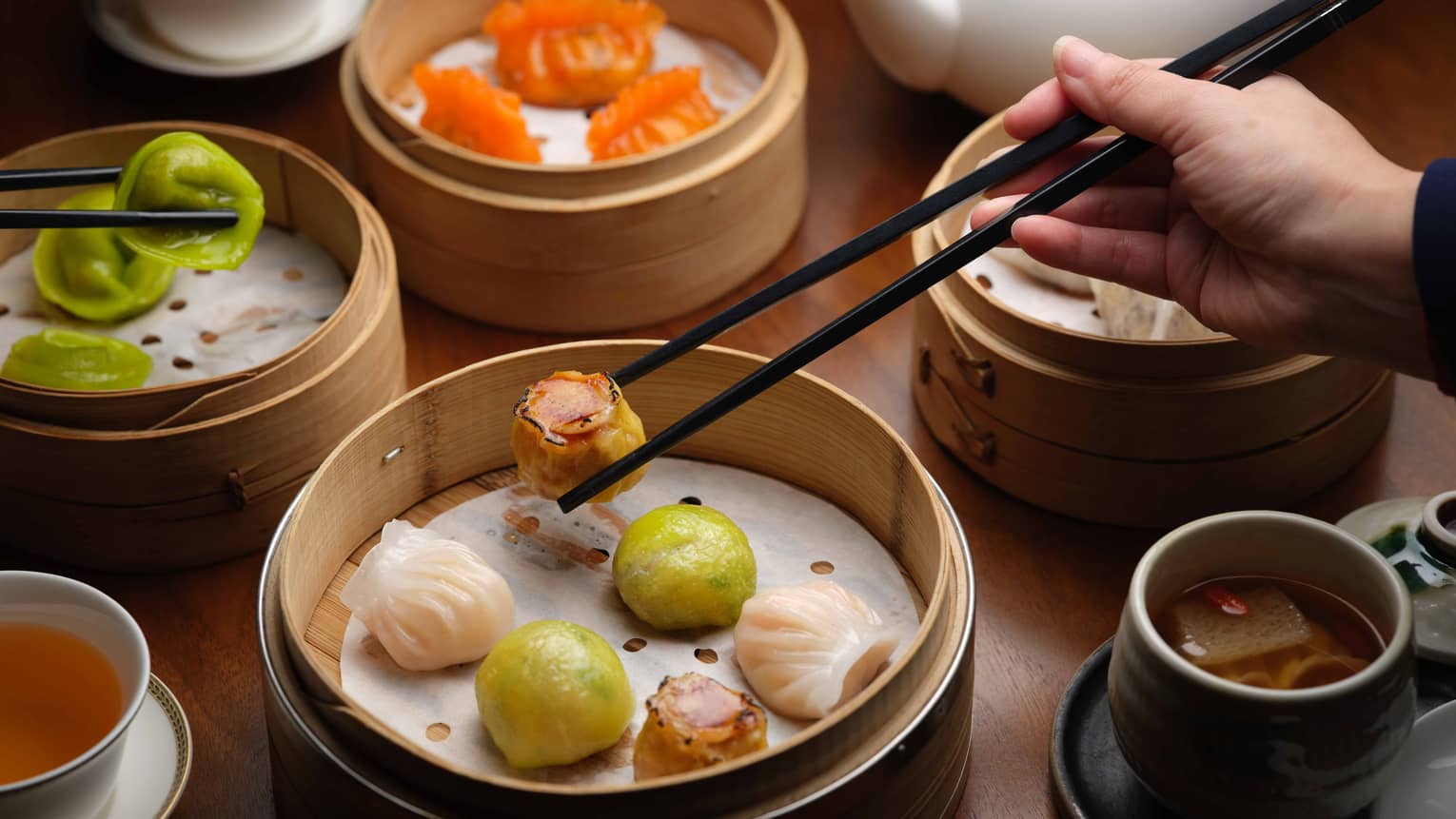 Beside cups of tea, a hand uses chopsticks to pick up one of the varied pieces of dim sum picked from nearby bamboo steamers.