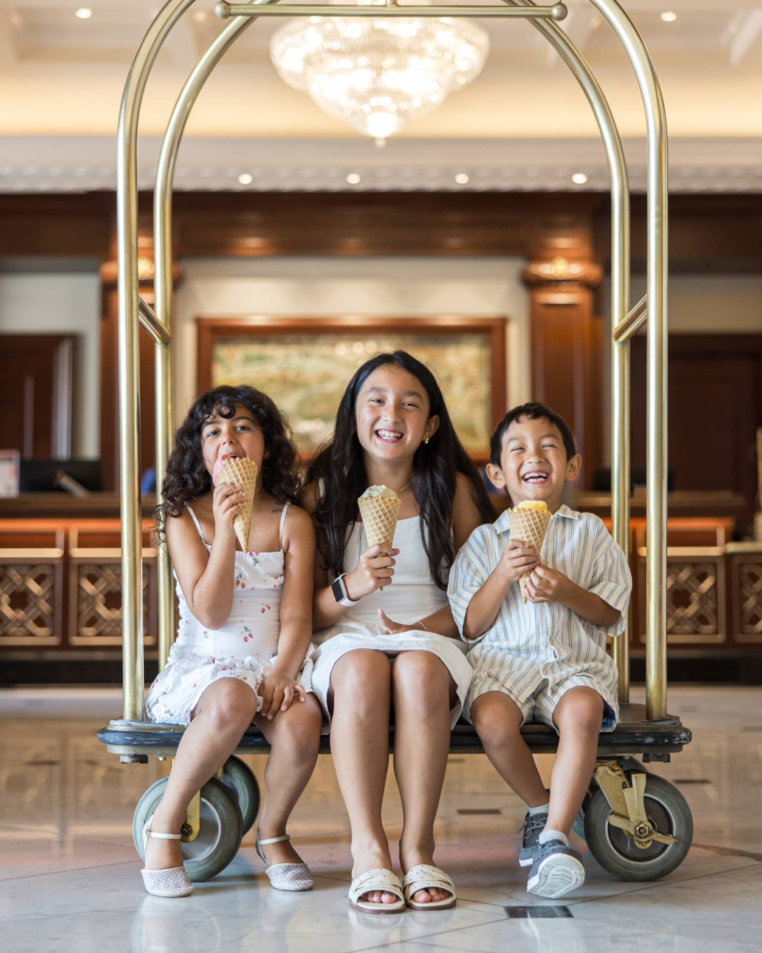 Three children sit on a gold hotel luggage cart eating ice cream