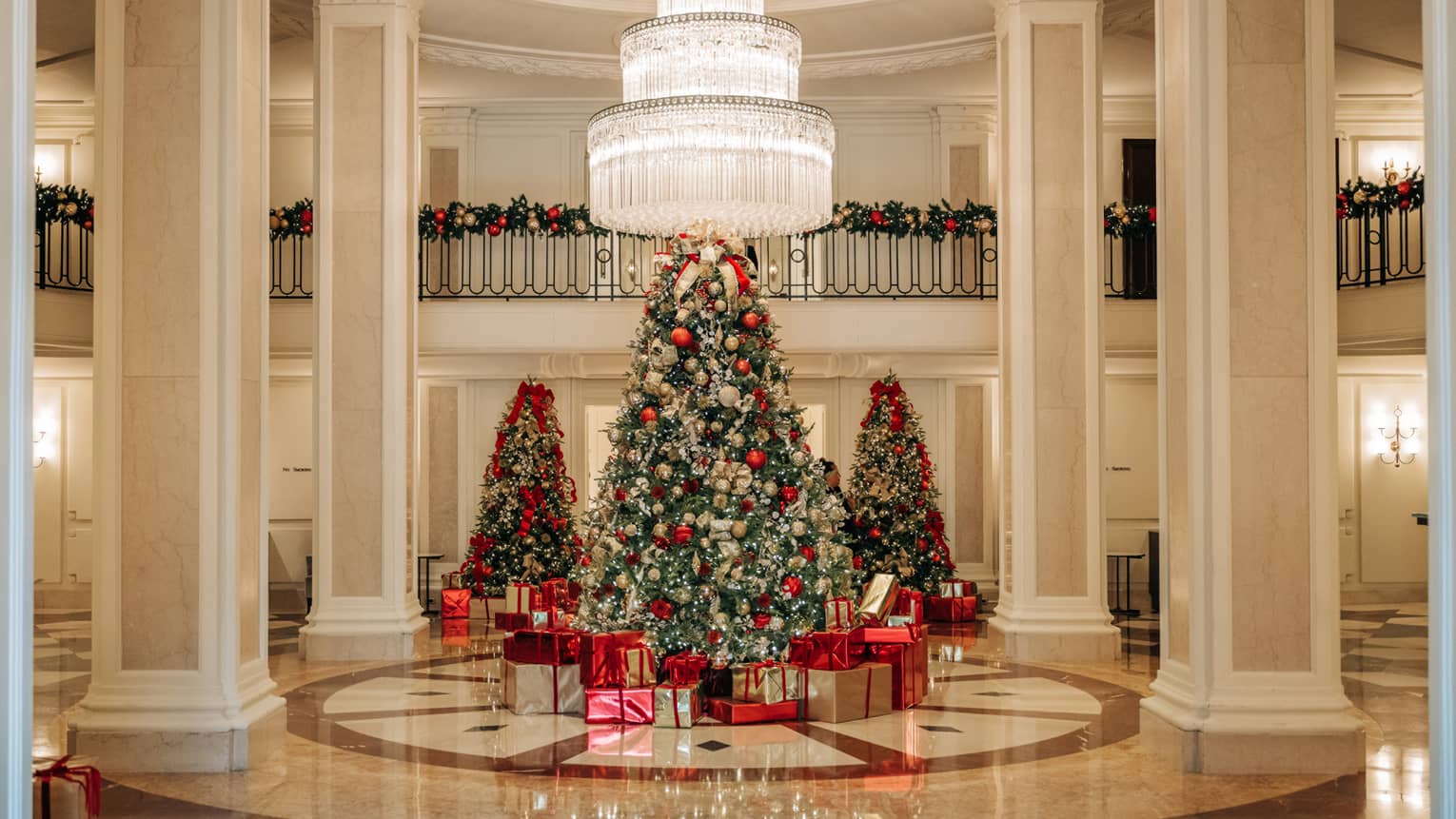 White marble hotel lobby decorated for Christmas with three trees each decorated in red, silver and gold and green garlands draped over the second floor balcony railing