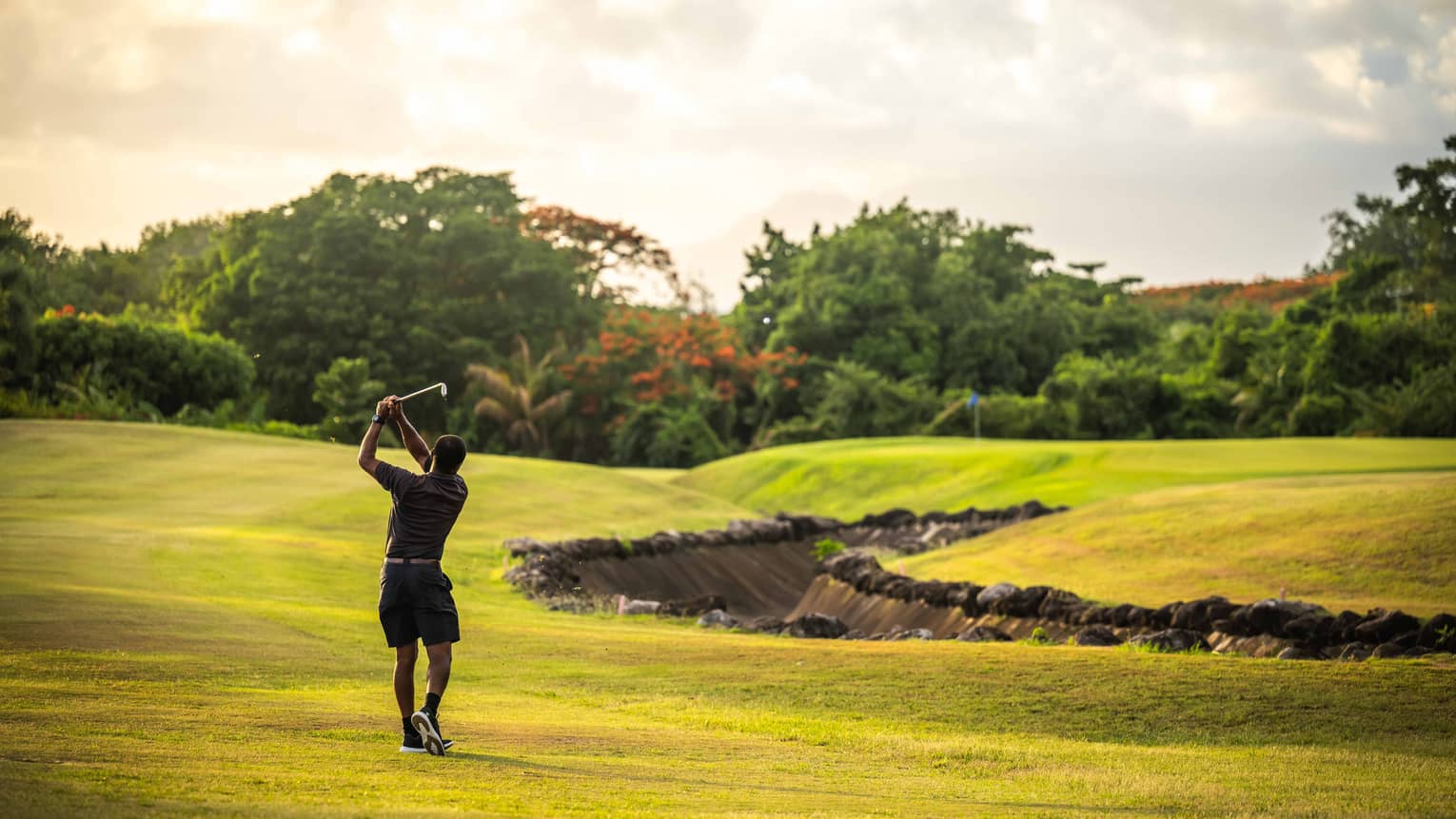 A golfer takes a swing on a bright green golf course surrounded by trees and lush vegetation, under a bright, partly cloudy sky