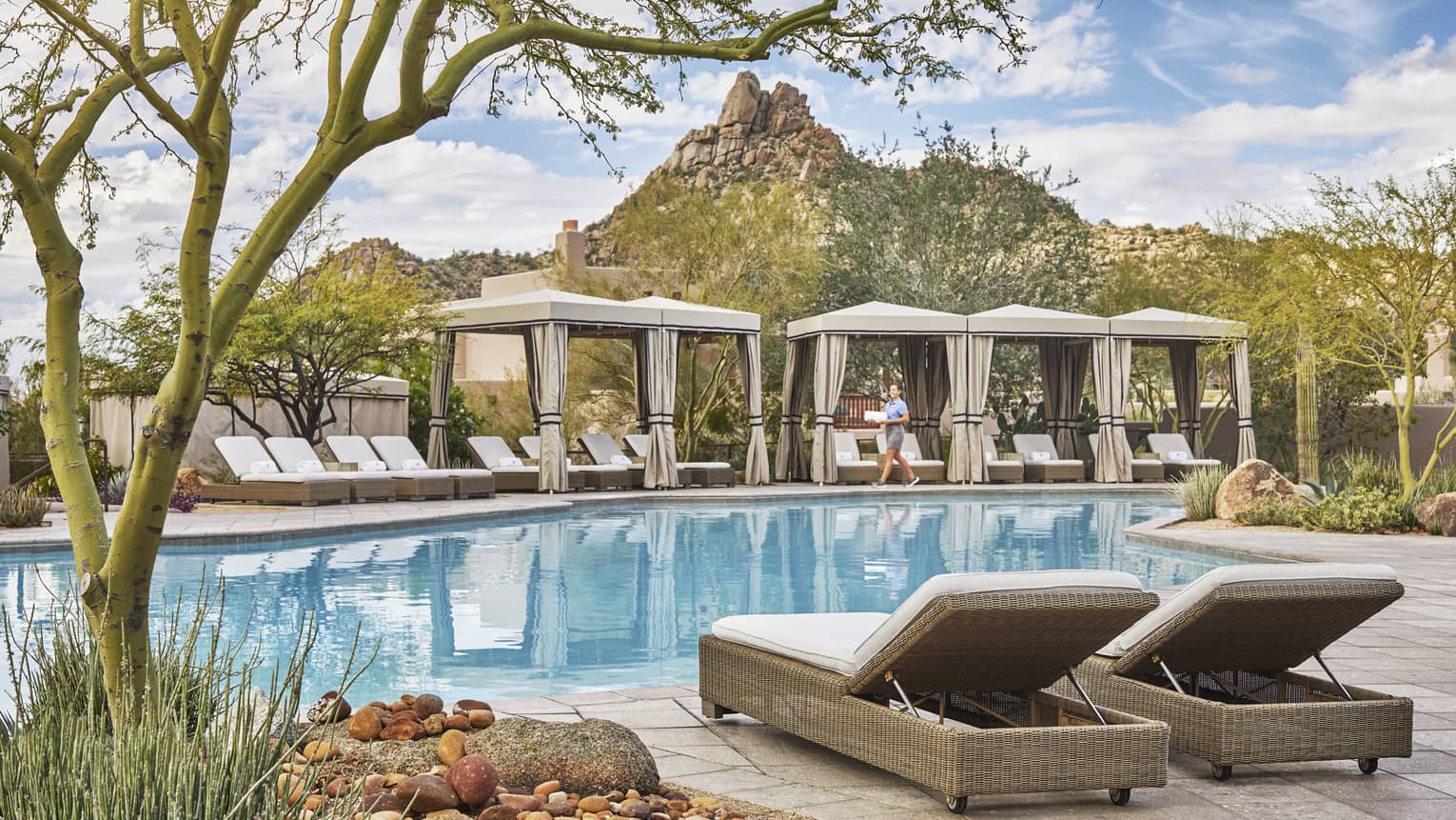 Outdoor pool with lounge chairs, canvas cabanas, staff member, rocky surroundings
