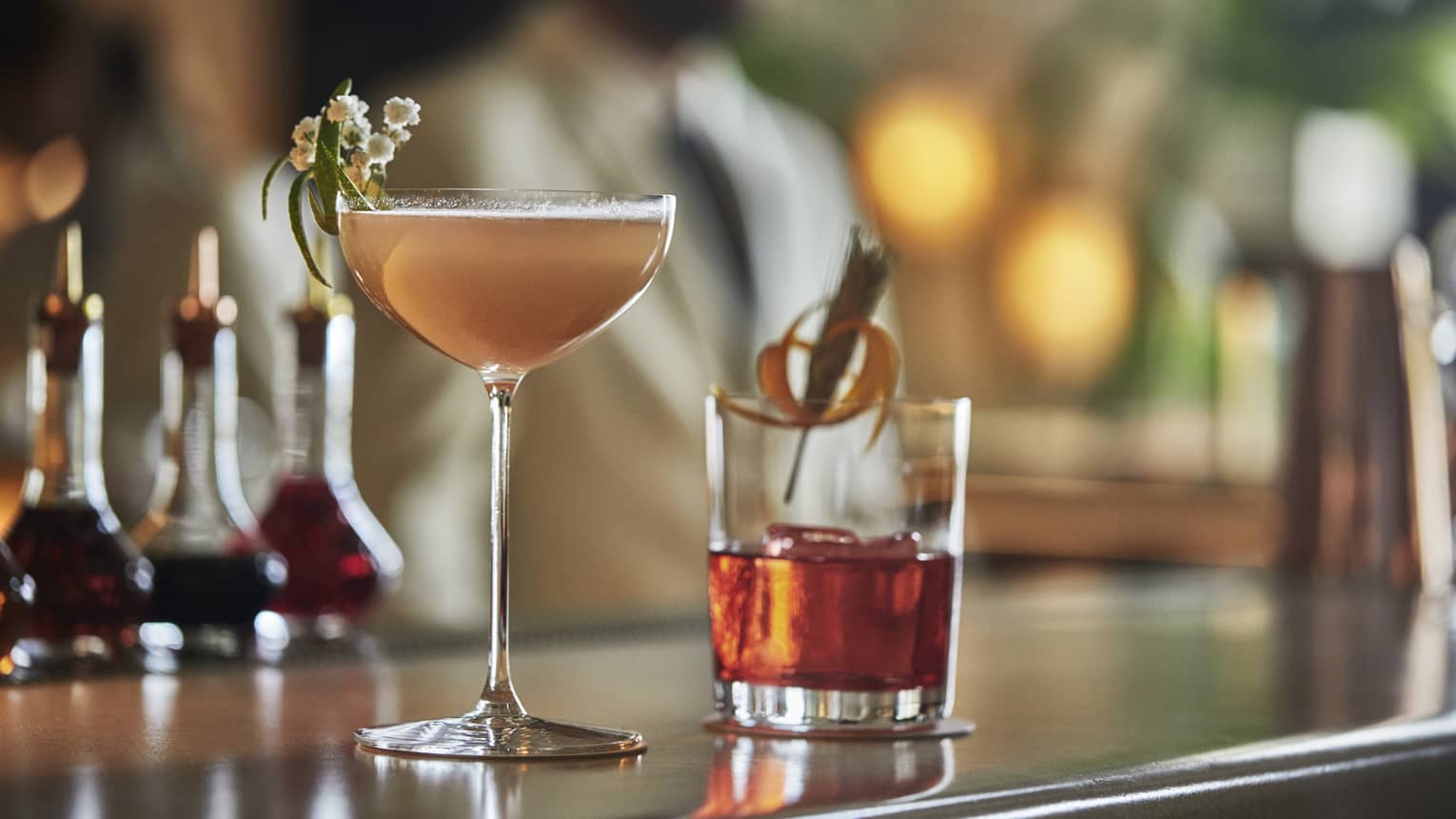 Two cocktails with garnish sit on a bar top with bottles and blurred image of man in background 