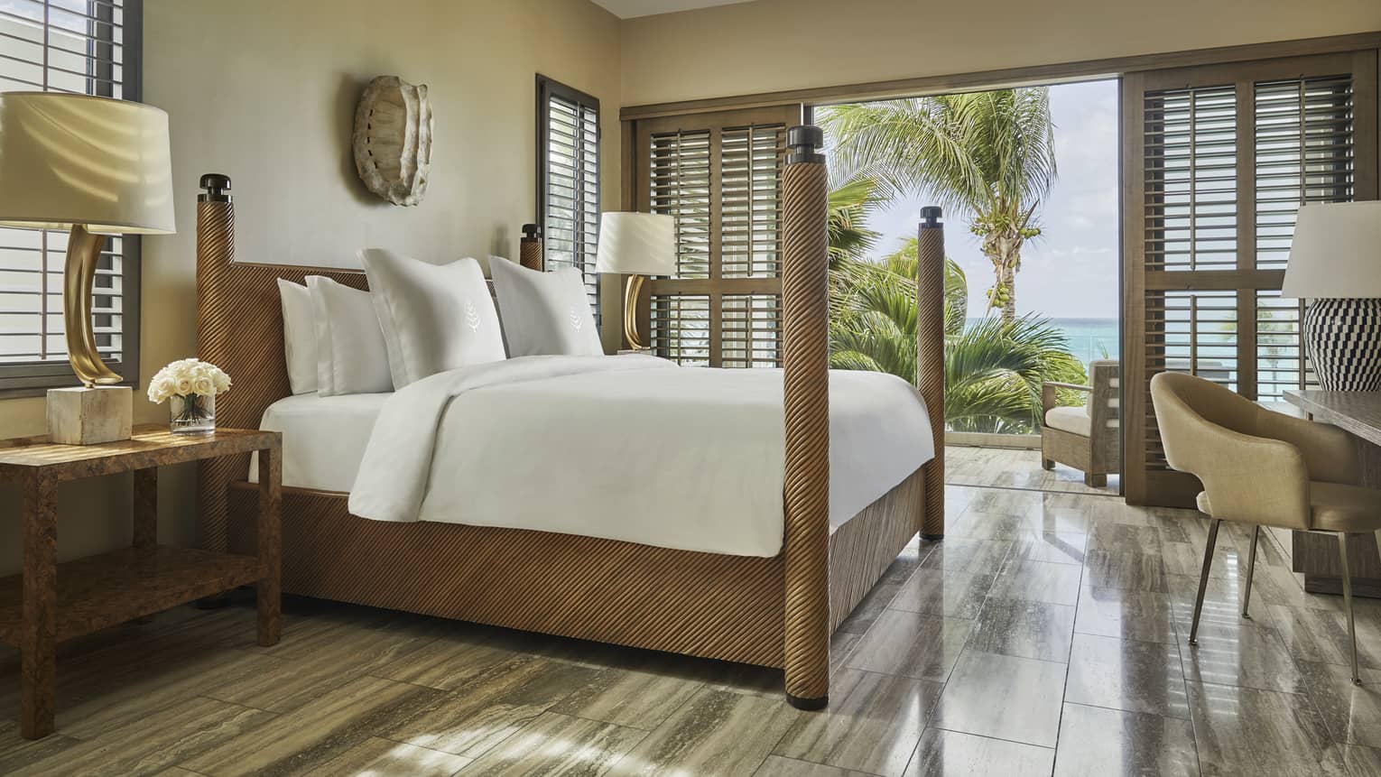 Luxury beachfront villa bedroom with a four-poster bed, white bedding and decorative lamps, featuring a balcony view of tropical palm trees and the ocean