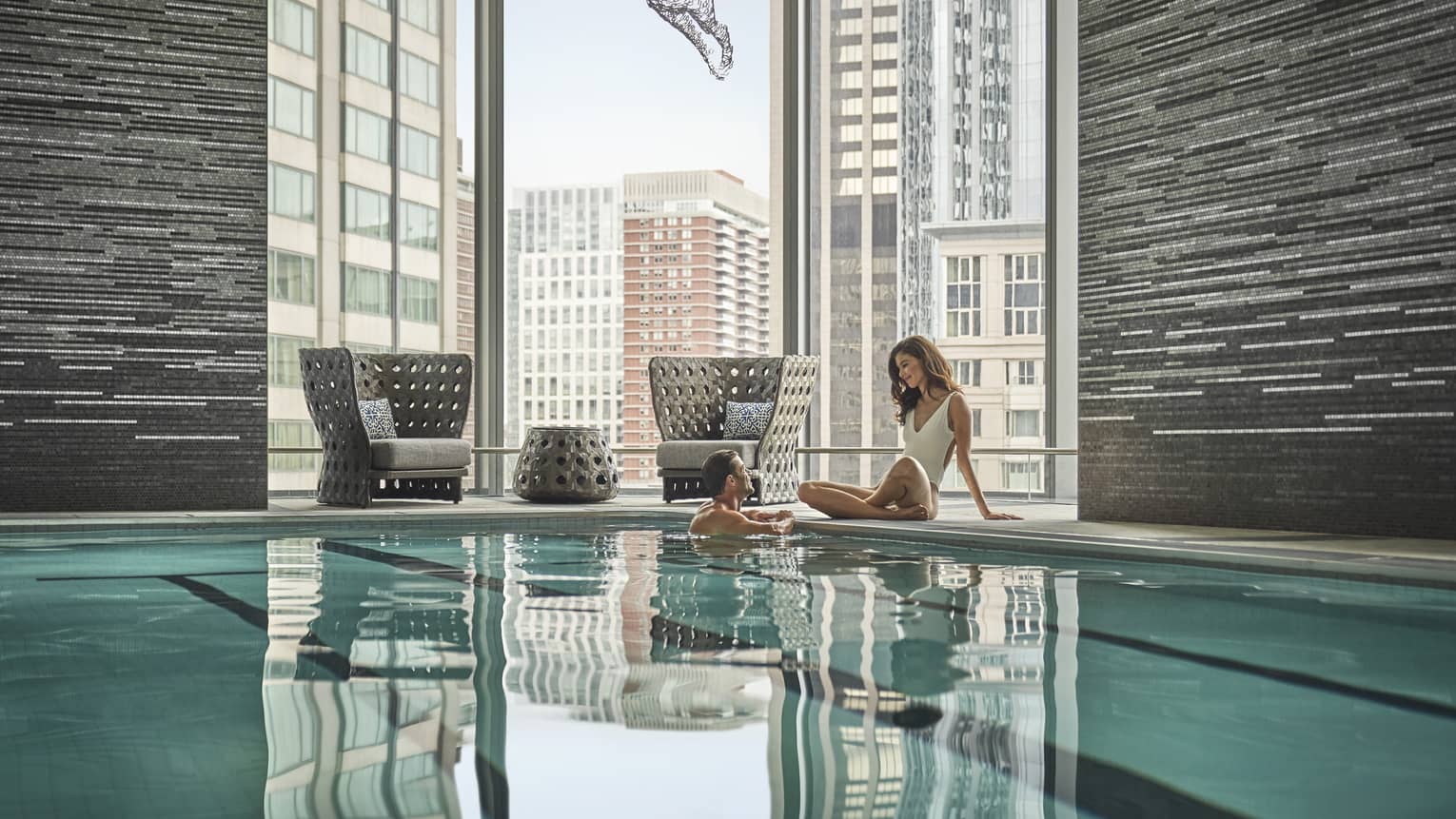 A couple embraces in a lap pool; behind them, floor-to-ceiling windows reveal an urban landscape.