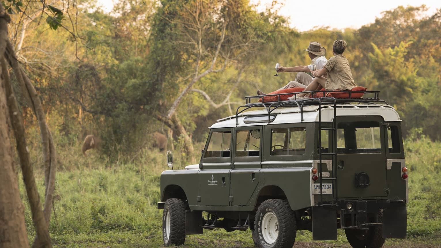 A couple sits on the roof of a Land Rover in the Bush, drinking a cocktail