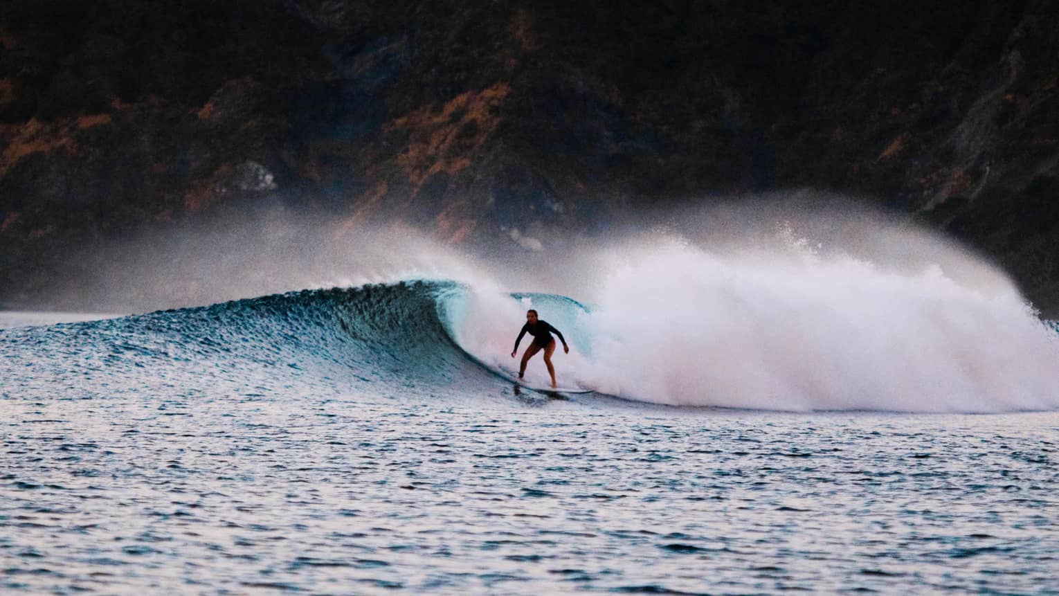 A surfer rides a wave amid a great cloud of frothy sea foam; a jagged cliff in the background, pink and yellow skies above.  