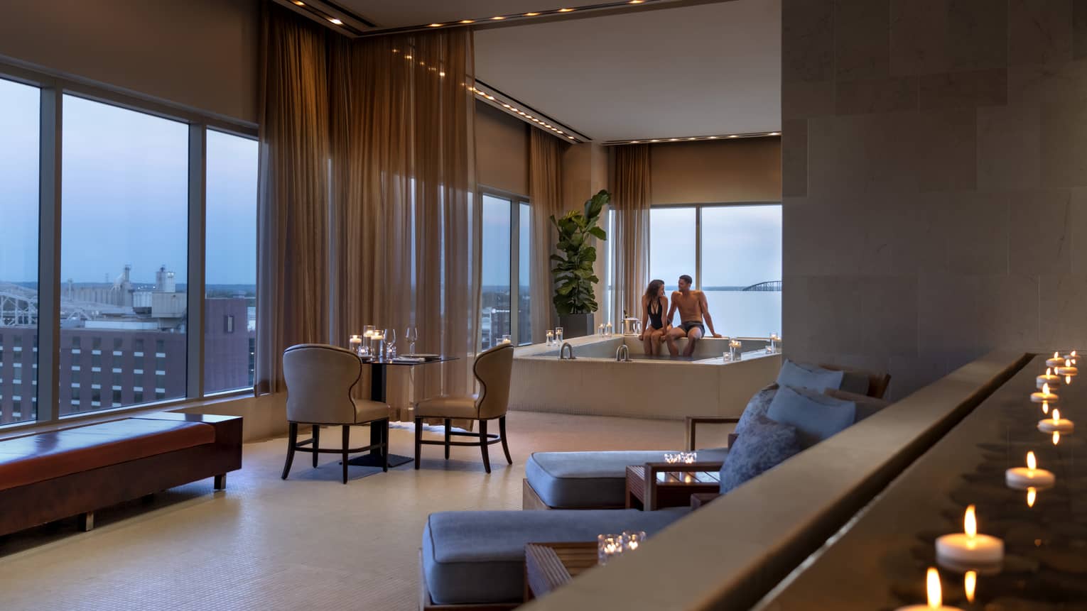 A couple enjoying a candlelit hot tub in a luxury hotel spa, with elegant seating, soft lighting and large windows offering a view of a distant bridge