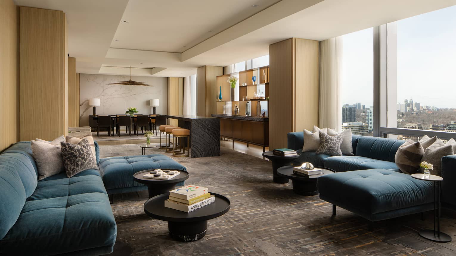 Luxury suite living area with blue sectional sofas, round coffee tables, bar with stools, dining table and floor-to-ceiling windows with city views