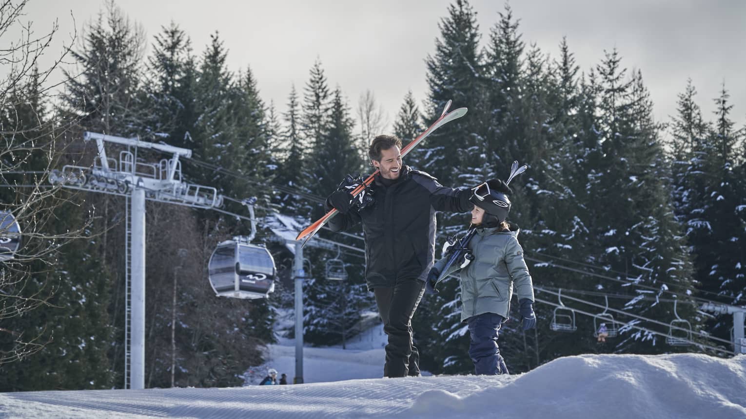 Smiling man and young son carry skis on their shoulders down the slope, ski lift in background 