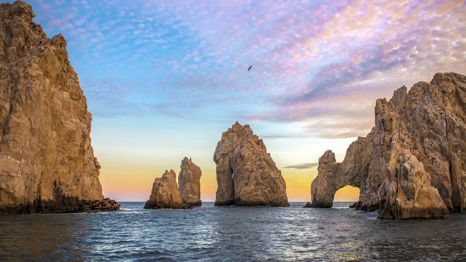 A bird soars through a multi-hued, dappled sunset sky. Towering sea stacks cast shadows over glistening reflective waters.