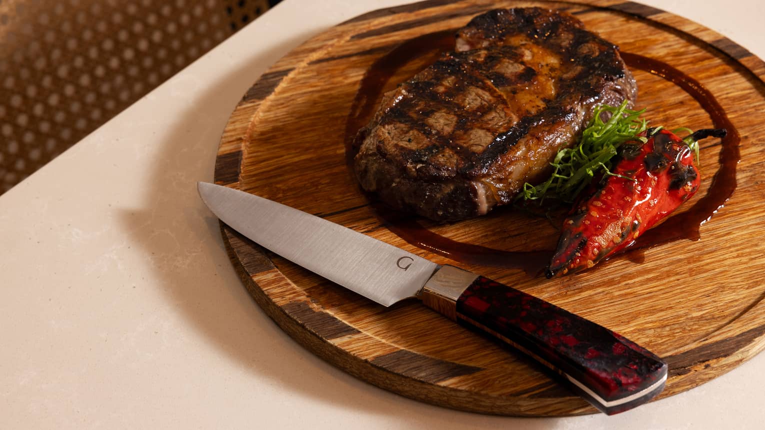 A round wooden serving plate with a piece of steak and garnishes. A steak knife sits on the plate near the edge.