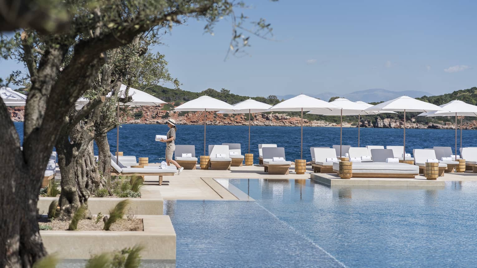 Woman holding towel walks past hotel pool with white chairs and umbrellas beside blue ocean