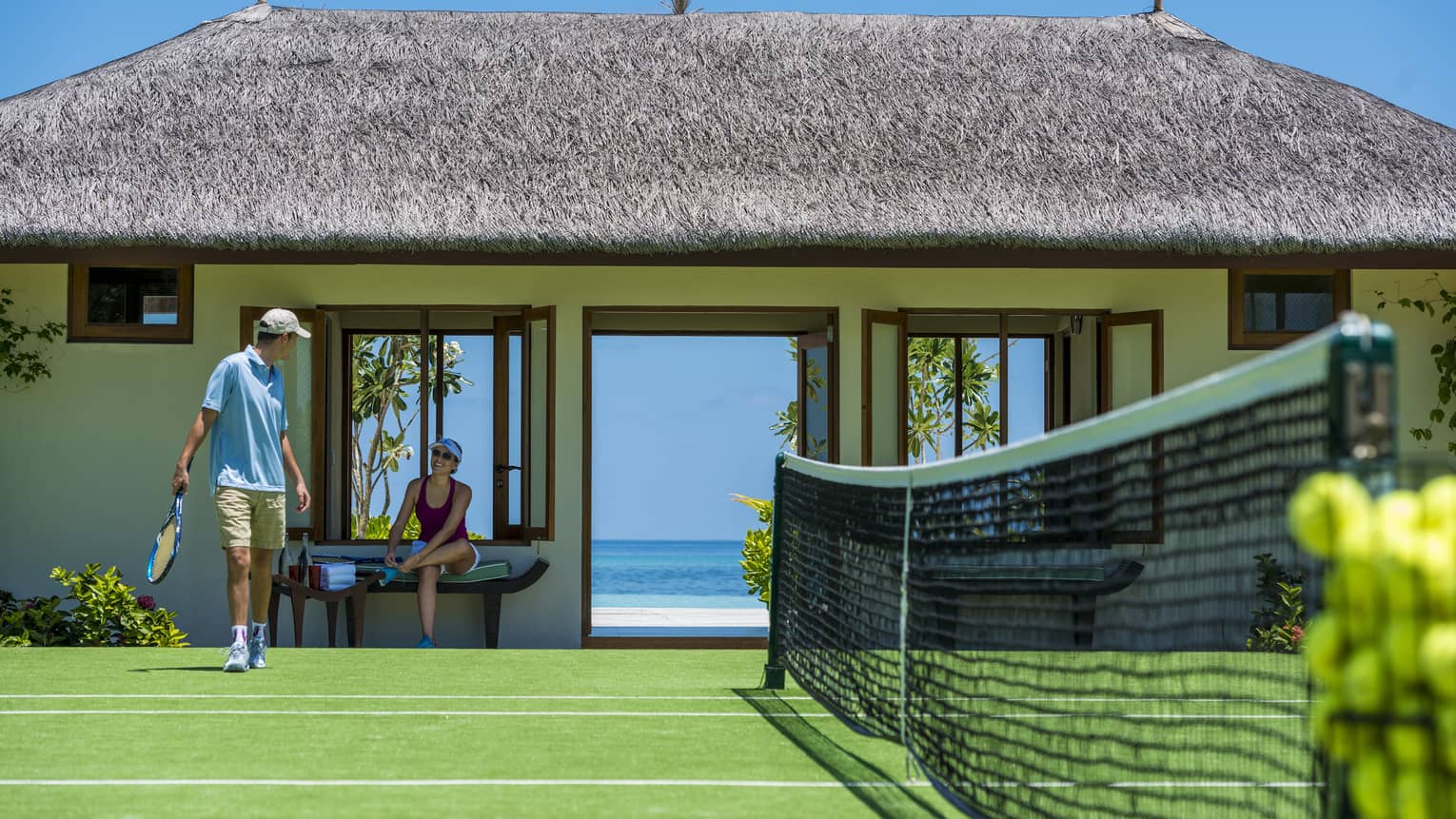 Two guests prepare to play tennis on a court bordered by a grass-roofed building with doors opening onto a view of the ocean.