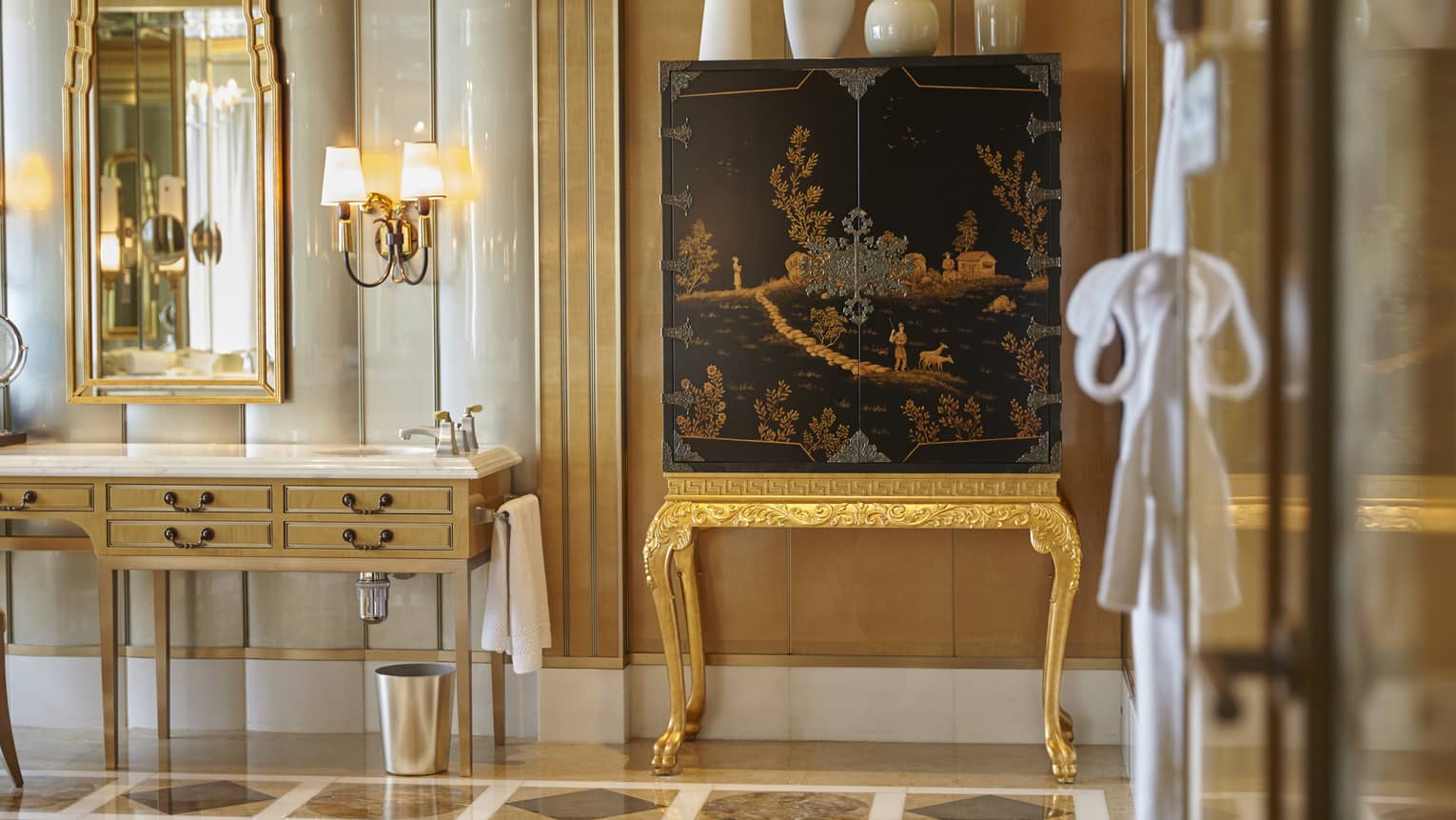 Elegant bathroom with a gold vanity, ornate mirror, and a black and gold decorative cabinet. A wall sconce and a hanging robe are also visible.
