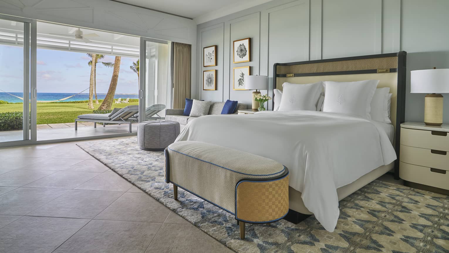 Luxury oceanfront bedroom with a large bed, cozy seating and sliding glass doors opening to a private patio with lounge chairs 