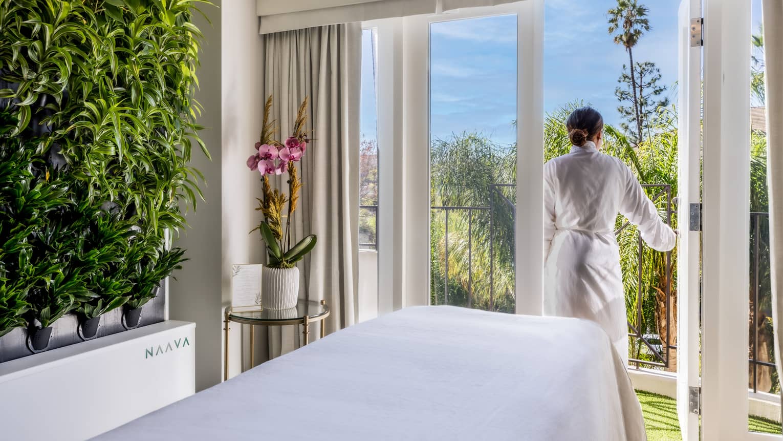 A woman walking outside from a massage room with large windowed doors and plants on one wall.