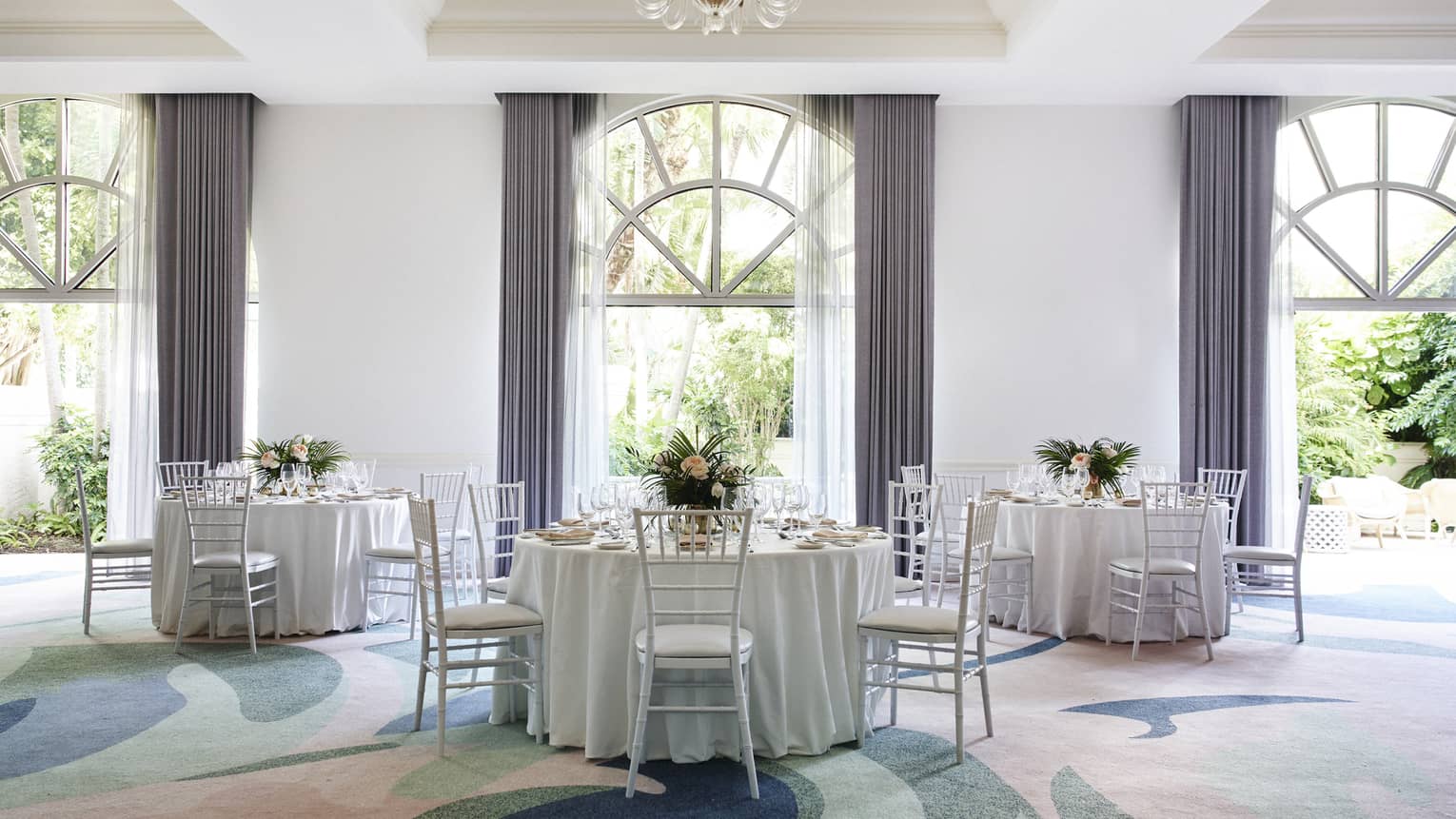 A large room with colourful carpet, round tables with white cloths, and large windows.