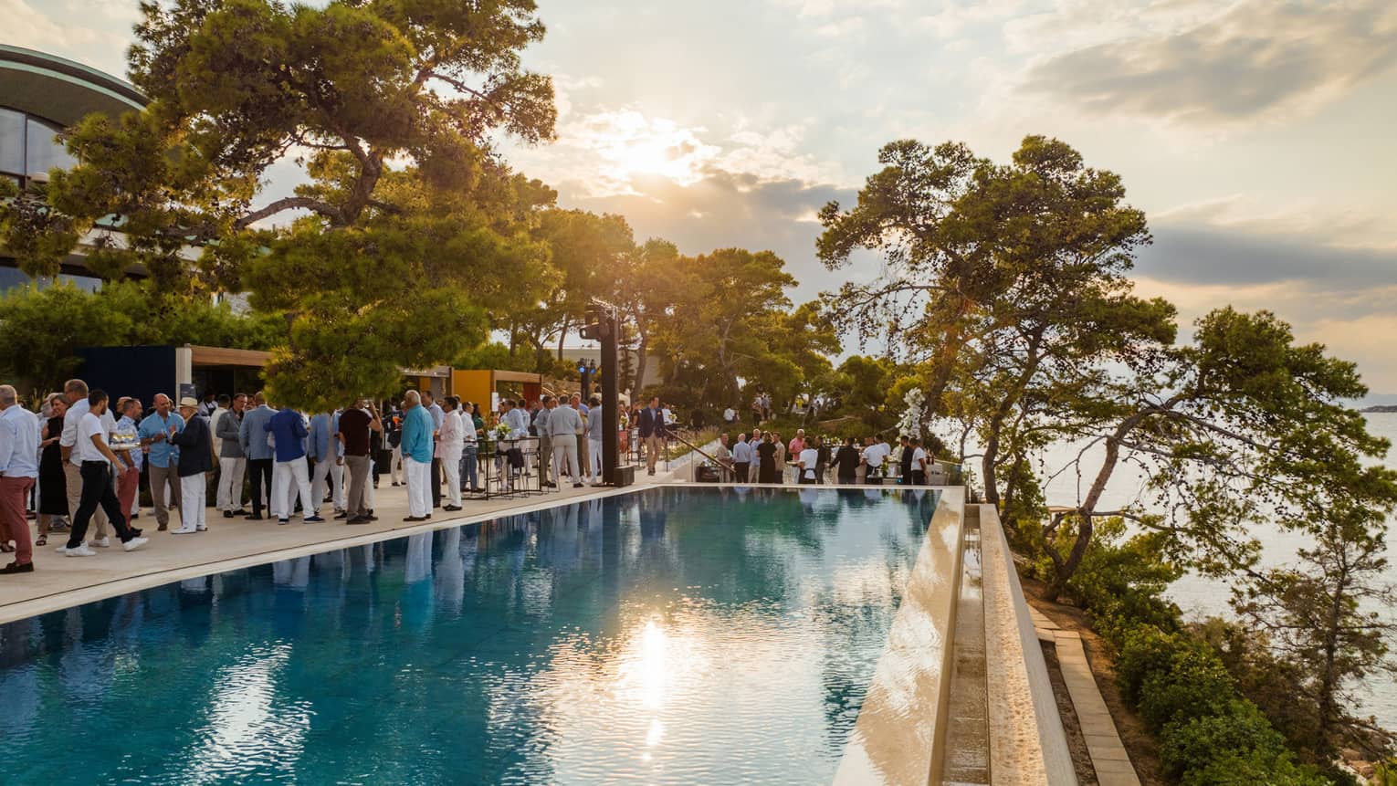 Group of people standing beside infinity pool with view of Saronic Gulf at sunset