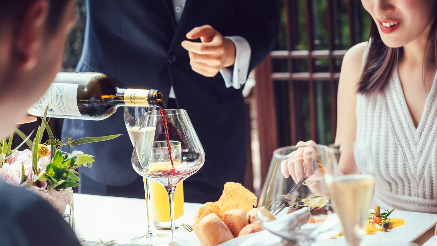 Server pouring red wine into large glass as couple dines at table