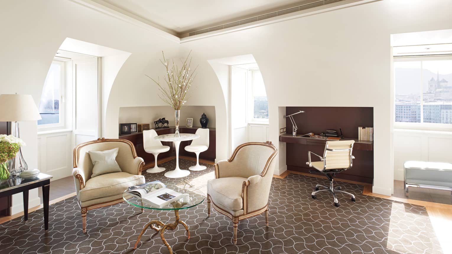 Four Seasons executive suite with antique-style armchairs, a glass coffee table, modern dining set, built-in desk and arched windows offering city views