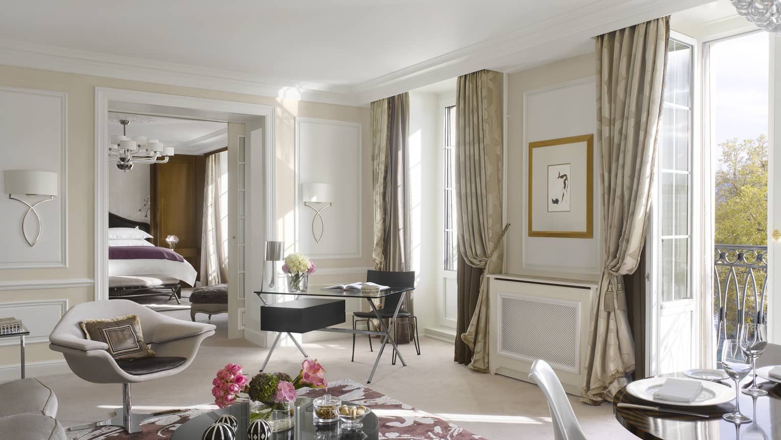 Luxury hotel suite living area with a modern armchair, black desk, round coffee table, dining table and balcony access, leading to a bedroom with a king bed