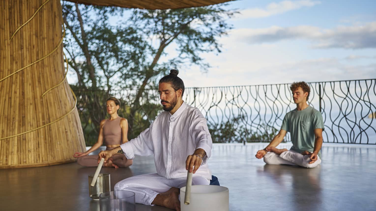 Man wearing all white clothing sits cross-legged on floor with three singing bowls in front of him and two people also sitting cross-legged on the floor behind him